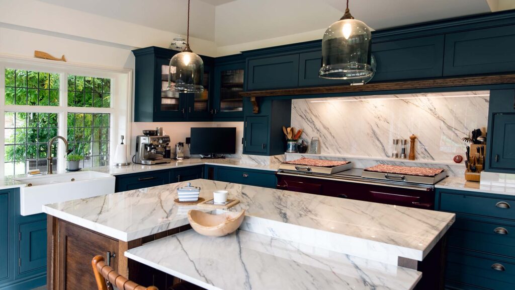 A featured image of a beautiful bespoke kitchen in Staplehurst, Kent. The space features classic green shaker-style cabinets, a large central island with a wooden countertop, and a rustic stone floor. The room is filled with natural light, and a bouquet of flowers sits on the countertop.