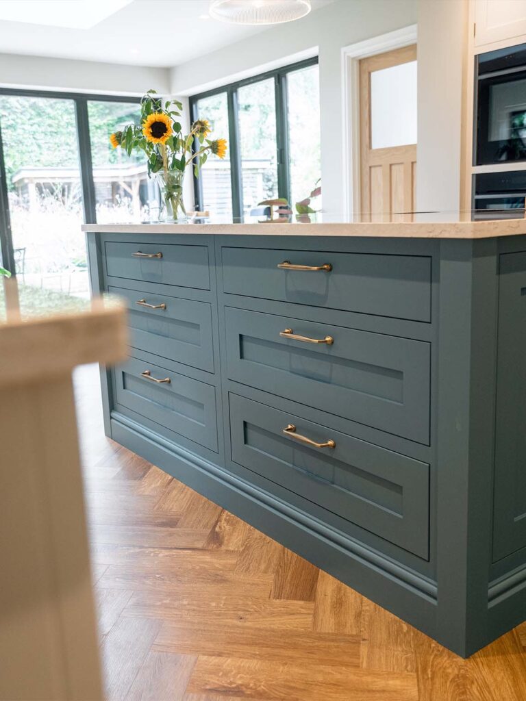 A wide shot of a bespoke kitchen showing the bright blue base cabinets and white wall cabinets. A large central island with a wooden breakfast bar and white stools sits in the foreground.