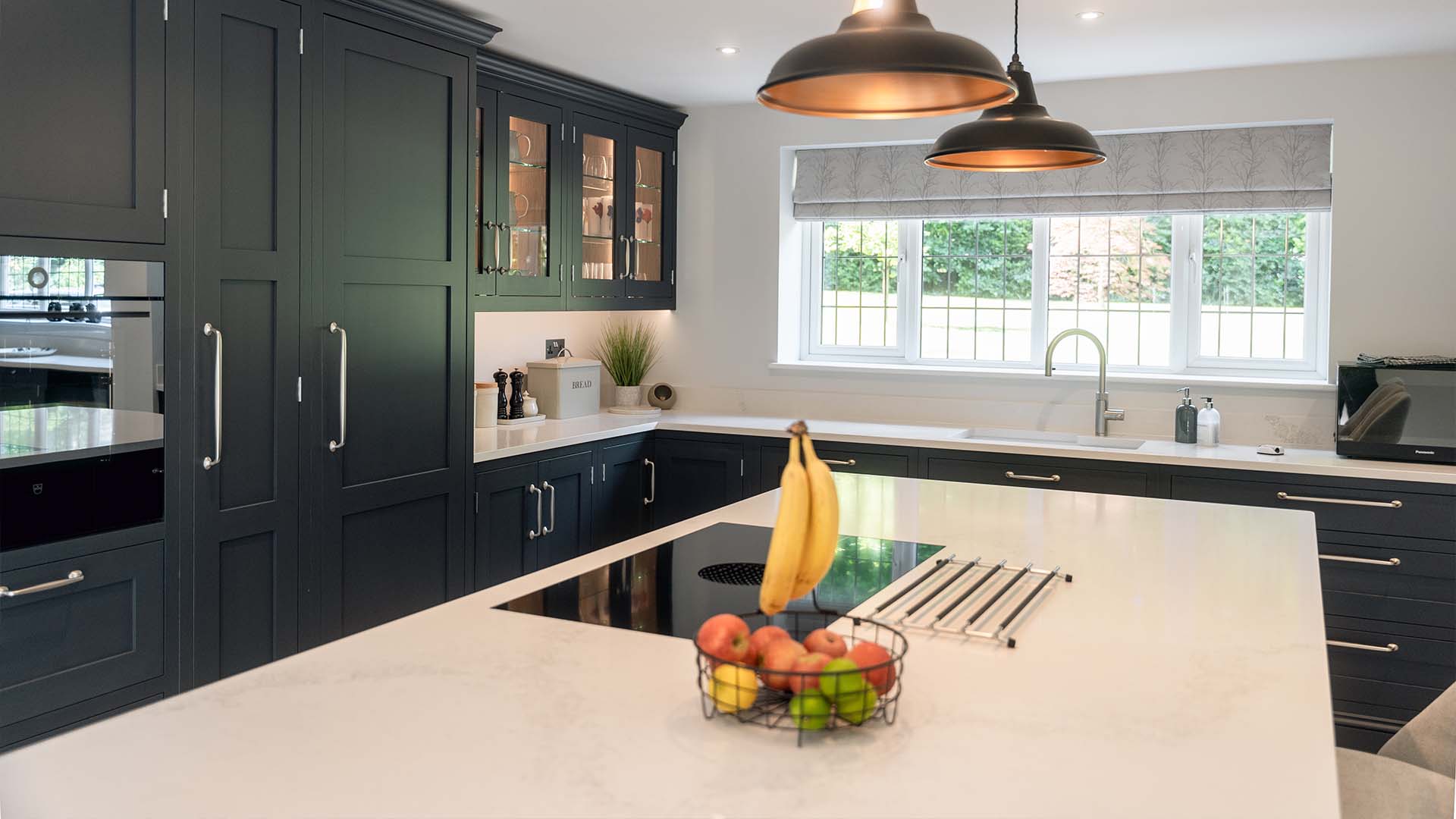 A wide shot of a bespoke kitchen with bright blue cabinets and a white pitched ceiling with exposed wooden beams. The room is filled with natural light from a large window and features a stylish pendant light over the island.