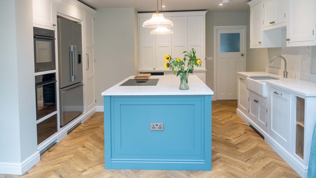A featured image of a vibrant bespoke kitchen in Crowborough, East Sussex. The space has bright blue, classic shaker-style cabinets, a white, patterned countertop, and a large central island. The room has a white, pitched ceiling with exposed wooden beams, and a minimalist pendant light hanging over the island.