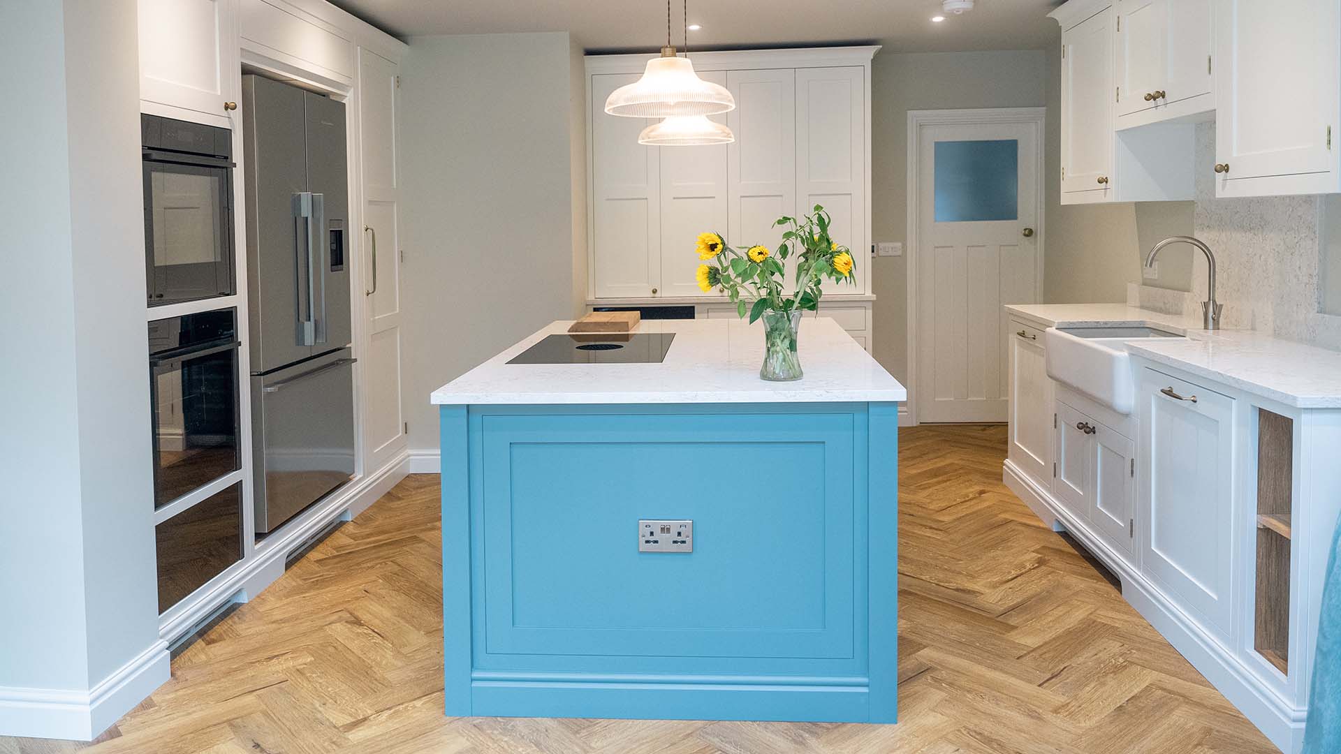 A featured image of a vibrant bespoke kitchen in Crowborough, East Sussex. The space has bright blue, classic shaker-style cabinets, a white, patterned countertop, and a large central island. The room has a white, pitched ceiling with exposed wooden beams, and a minimalist pendant light hanging over the island.